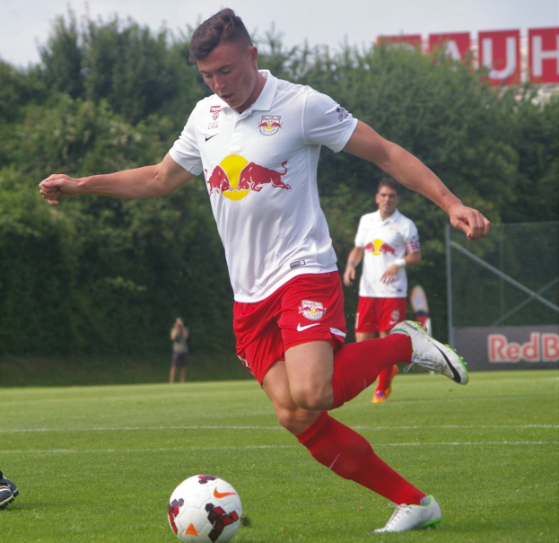 Professioneller Fußballspieler, der einen Ball auf einem grünen Feld mit dem 'RB Leipzig'-Schild im Hintergrund unter einem klaren blauen Himmel tritt.