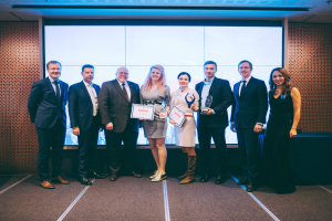 Gruppe von strahlenden Menschen mit Urkunden, die vor einem Bildschirm mit einem Tisch und einem Objekt im Hintergrund stehen, Gewinner der Internationalen Business Awards 2018.