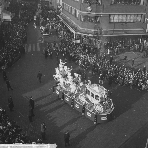 Eine große Menschenmenge umringt einen Paradewagen, der mit bunten Ballons und Bändern geschmückt ist, mit Gebäuden und Laternen im Hintergrund, in Schwarz-Weiß.