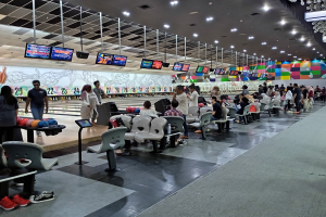 Eine Gruppe von Menschen sitzt in einer Bowlinghalle mit Bowlingkugeln auf den Tischen, einige stehen links, einer hält eine Kugel rechts, unter einer Decke mit Lampen und Fernsehern.