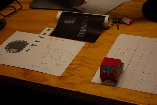 Ein Tisch in der Mitte mit einem Papier, einer Grafik und einigen Schlüsseln daneben, Drähte oben links und ein Spielzeugauto unten rechts.