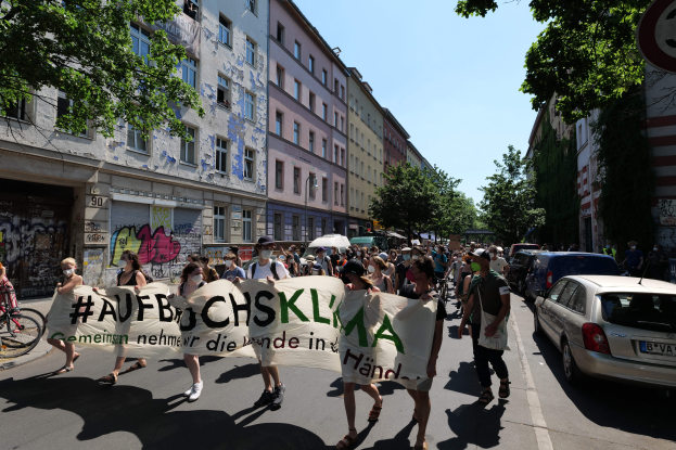 Eine Gruppe von Menschen marschiert auf einer Straße in Berlin, hält eine Fahne hoch, mit parkenden Autos auf der rechten Seite, einem Radfahrer auf der linken Seite und Gebäuden, Bäumen und einem Schild im Hintergrund unter einem sichtbaren Himmel.