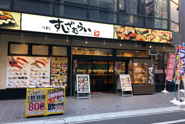 Außenansicht einer Sushi-Bar in Tokyo mit Glaswänden, Essensauslagen, Schildern und einer Straße im Vordergrund.