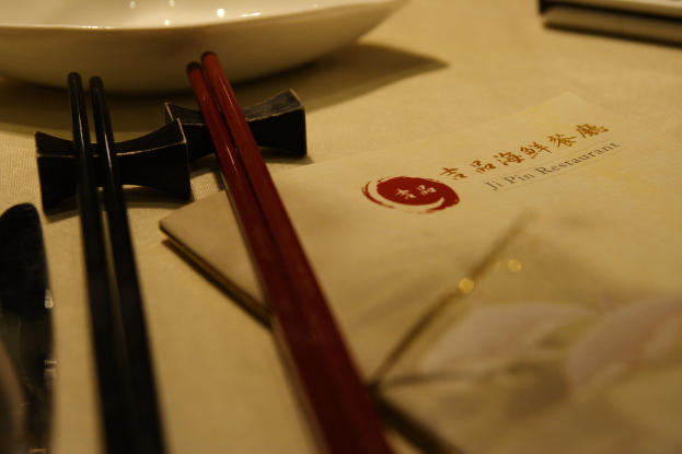 Ein Tisch mit einer Speisekarte, die auf Mandarin und Englisch 'Ji Pin Restaurant' heiÃt, sowie zwei Paar StÃ¤bchen (eines rot, eines schwarz) und eine weiÃe SchÃ¶Ãel davor.