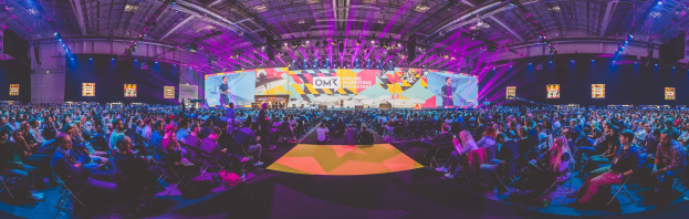 Eine große Menschenmenge, die auf Stühlen sitzt und einer hell erleuchteten Bühne mit mehreren Personen darauf gegenübersitzt, umgeben von Bildschirmen, wahrscheinlich auf einem Online-Marketing-Rockstars-Event.