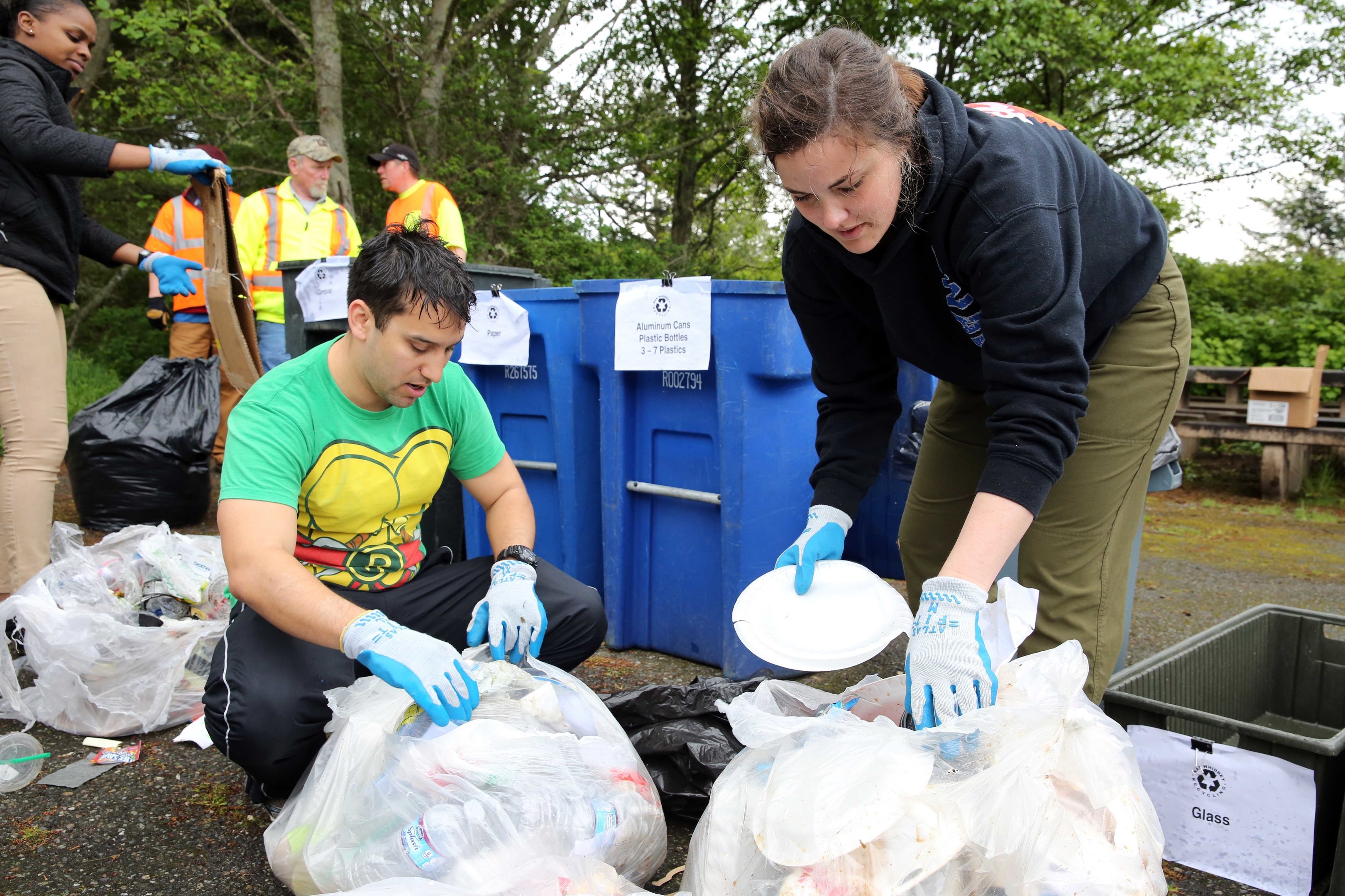 Eine Gruppe von Menschen, die Müll in einem Park sammeln, mit zwei Personen in der Mitte, die Handschuhe tragen und Schilder halten, umgeben von weggeworfenen Gegenständen, einem Mülleimer, einer hölzernen Bank, Bäumen und einem klaren blauen Himmel.