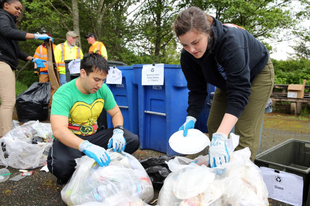 Eine Gruppe von Menschen, die Müll in einem Park sammeln, mit zwei Personen in der Mitte, die Handschuhe tragen und Schilder halten, umgeben von weggeworfenen Gegenständen, einem Mülleimer, einer hölzernen Bank, Bäumen und einem klaren blauen Himmel.