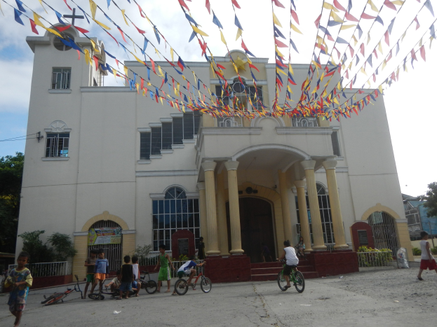 Gruppe von Menschen auf Fahrrädern vor einer weißen Kirche mit architektonischen Details, umgeben von Grün, einem Banner, Bäumen und einem bewölkten Himmel, mit bunten Fahnen darüber.
