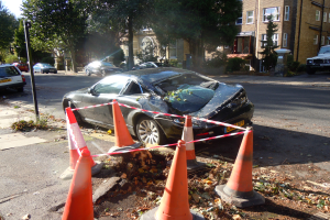Ein verunglücktes Auto am Straßenrand, umgeben von orangen Verkehrskegeln, mit Bäumen, Gebäuden mit Fenstern und einem klaren blauen Himmel im Hintergrund.