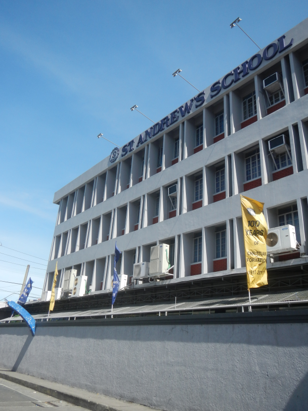 Ein Schulgebäude mit Fenstern, Klimaanlagen, Flaggen, einem Namensschild, Straßenlaternen, Strommasten, Stromkabeln und einer bewölkten Himmel im Hintergrund.