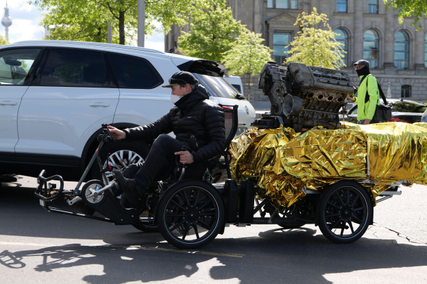 Ein Mann im Rollstuhl mit einem großen Motor auf dem Rücken, umgeben von Fahrzeugen auf einer Straße, mit Bäumen, Gebäuden, Polen und einem klaren blauen Himmel im Hintergrund; er trägt eine schwarze Jacke, eine Mütze und hält ein Objekt in der Hand.