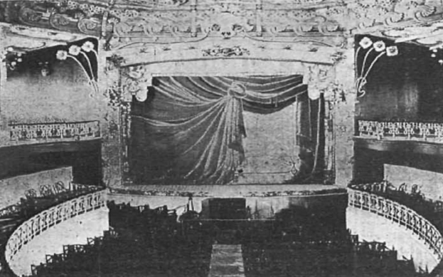 Schwarzes und weißes Foto des Auditoriums des Theatre Royal in London, das eine Bühne mit Vorhang, Geländern und ornamentalen Wanddesigns zeigt, sowie einige sitzende Besucher und Text am unteren Rand.