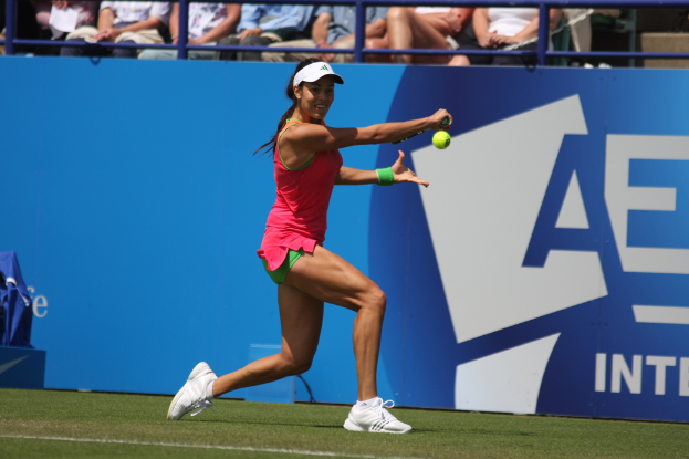 Eine Frau spielt Tennis, hält einen Schläger in ihrer rechten Hand, trägt eine weiße Mütze und Schuhe, mit einem grünen Band an ihrem linken Handgelenk, auf einem grünen Platz, mit Zuschauern im Hintergrund.