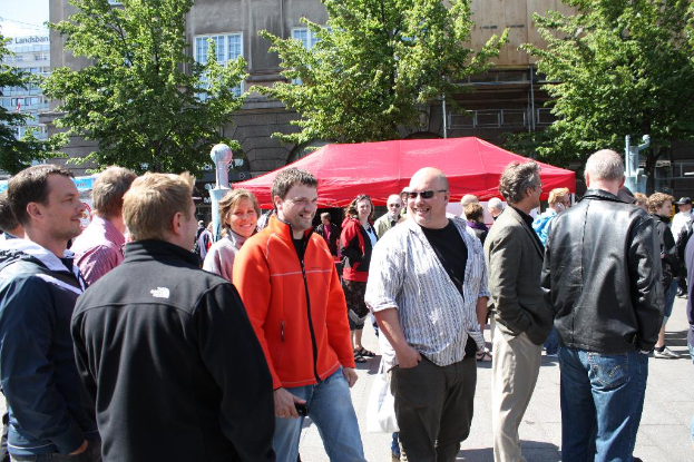 Eine belebte Straße mit vielen Menschen, ein Zelt, Bäume, Gebäude und Text auf einem der Gebäude.