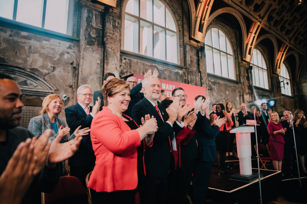 Eine Gruppe von Menschen, die vor einem Publikum stehen und applaudieren, näher bei einem Podium mit einem Mikrofon, einem Banner mit der Aufschrift "Covid-19 New York City Mayor Bill de Blasio" und Hintergrundelementen wie Stühlen, einer Wand und Fenstern.