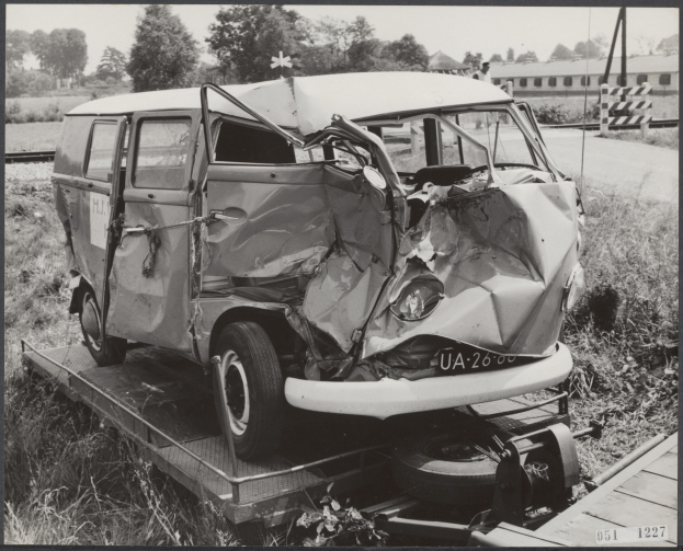 Ein Schwarz-Weiß-Foto eines schwerbeschädigten Autos auf einem Tieflader, umgeben von Gras und Pflanzen, mit Bäumen und einem Gebäude im Hintergrund.