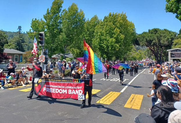Eine Gruppe von Menschen geht während des San Francisco LGBT Pride Parade die Straße entlang, einige halten Schirme, andere sitzen auf Stühlen oder stehen, mit einer Regenbogenflagge, einem Verkehrssignal, einem Schild, Bäumen, Gebäuden und dem Himmel im Hintergrund.