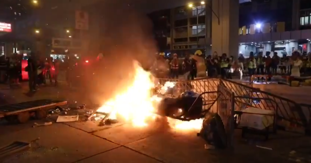 Eine Gruppe von Menschen steht um ein Feuer in der Mitte einer Straße, umgeben von Barrikaden und anderen Gegenständen, mit Gebäuden, Laternenmasten und einer Tafel mit Text im Hintergrund unter dem Nachthimmel.