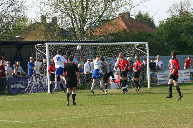 Fußballspieler sind auf einem Feld in einem Spiel involviert, mit einem Tor und Zuschauern im Hintergrund und Bäumen und Häusern weiter hinten.