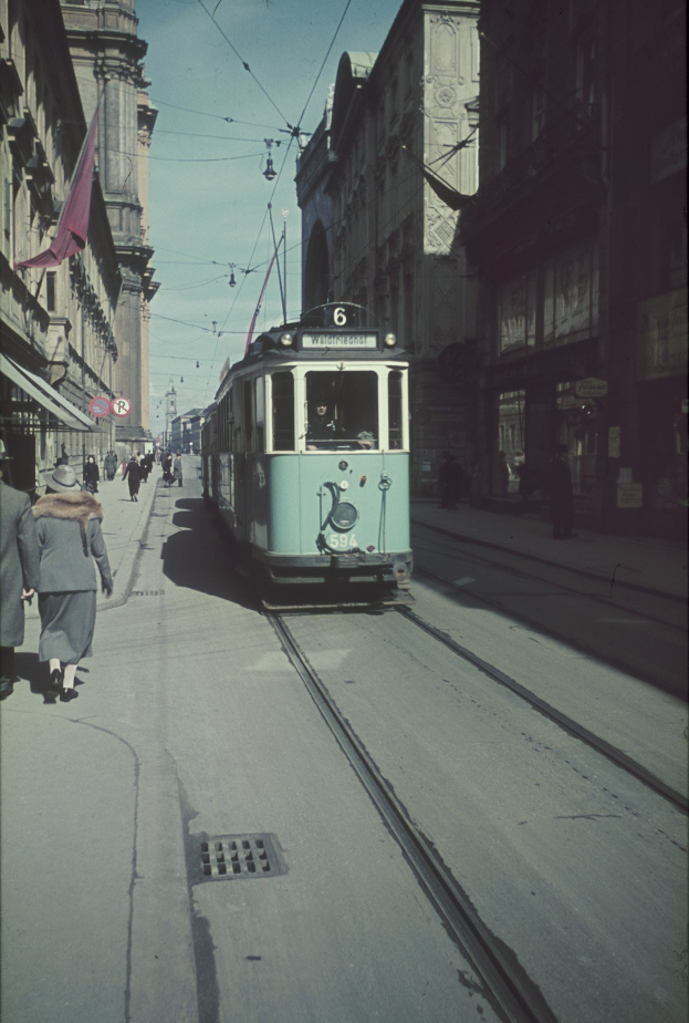 Altes Schwarz-Weiß-Foto einer Straßenbahn, die auf einer Stadtstraße mit Fußgängern auf dem linken Gehweg, Gebäuden, Strommasten und Drähnen im Hintergrund unter einem klaren Himmel fährt.