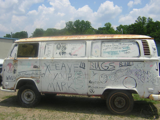 Ein beschädigtes Wohnmobil mit Gemälden und Zitaten darauf, das vor einer Gruppe von Bäumen geparkt ist.