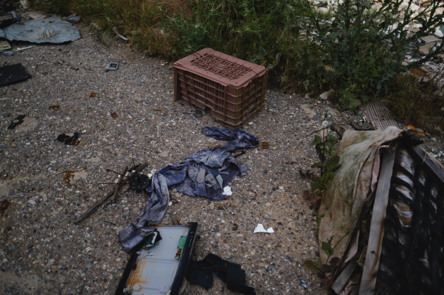 Haufen Müll einschließlich eines Plastikbehälters und Kleidung, die auf dem Boden neben einem Zaun verstreut sind, mit Pflanzen und Gras im Hintergrund.