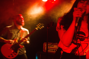 Eine Frau singt in ein Mikrofon auf der Bühne, während ein Mann im Hintergrund Gitarre spielt, mit Deckenlampen, die die lebendige Szenerie beleuchten.