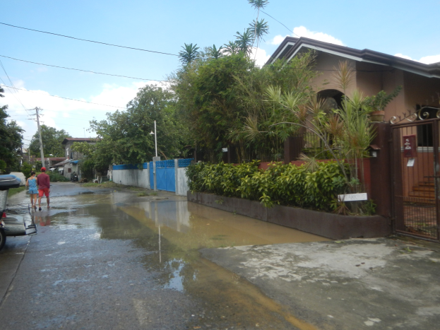 Eine überflutete Straße mit einem geparkten Motorrad, Menschen, die gehen, Gebäude, Bäume, Pflanzen, Pfähle, Drähte und ein Tor im Hintergrund unter einem bewölkten Himmel, mit einem Wasserzeichen auf dem Bild.