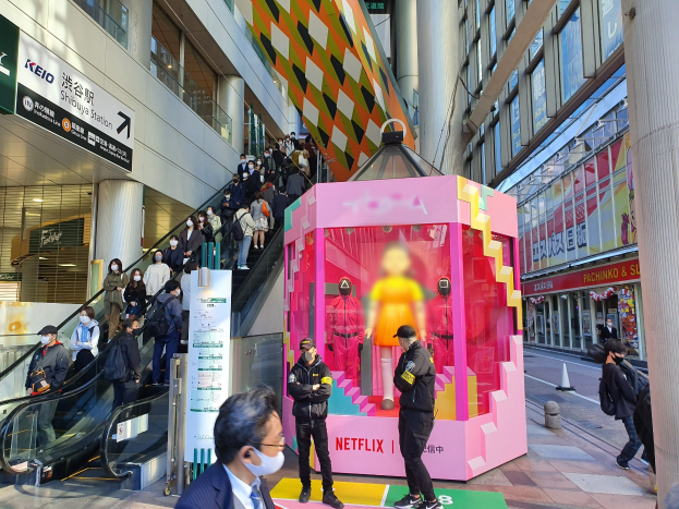 Eine Gruppe von Menschen steht vor einem Gebäude mit Glaswänden und Texttafeln, einige tragen Masken, während eine Rolltreppe mit Fahrgästen zu sehen ist; eine pinkfarbene Box in der Mitte zeigt Schaufensterpuppen und trägt die Aufschrift "Netflix's new Netflix booth in Tokyo."