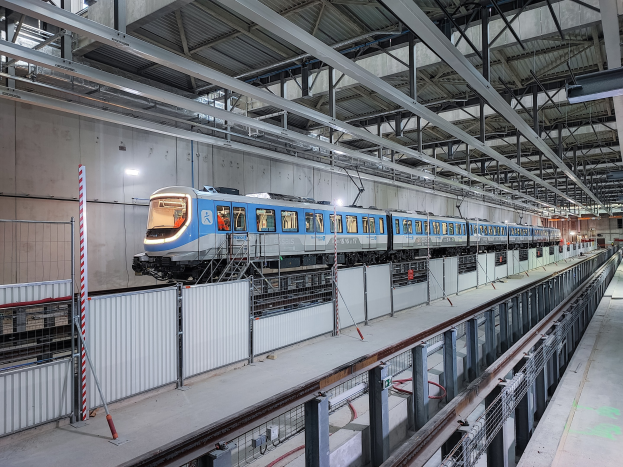 Blauer und weißer Zug fährt durch eine große Bahnhofshalle mit einem Bahnsteig auf der rechten Seite und einer Wand im Hintergrund, unter einer Decke mit Stangen und Lampen.