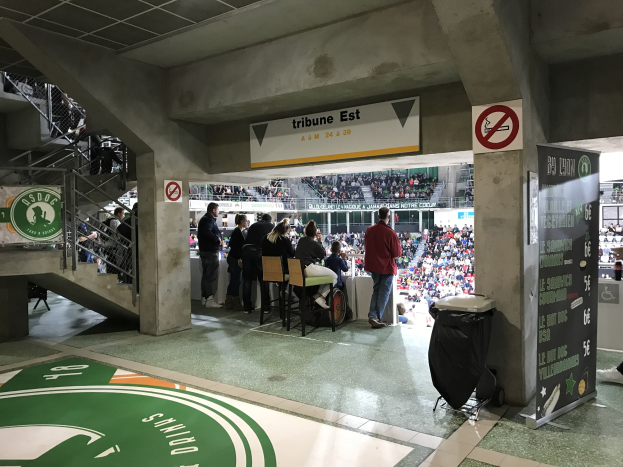 Eine Gruppe von Menschen steht vor einer sitzenden Menge in einem Stadion, mit Schildern, die "Boston Celtics vs New York Jets" anzeigen, einem Müllcontainer, einer Tafel, einer Treppe mit Geländern und einer Decke mit Deckenlampen.