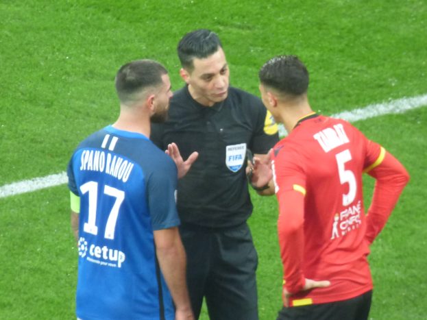Ein Schiedsrichter in schwarzer Kleidung spricht mit zwei Männern auf einem Fußballfeld, einer in blauen und roten T-Shirts mit Text, beide tragen eine Brille.
