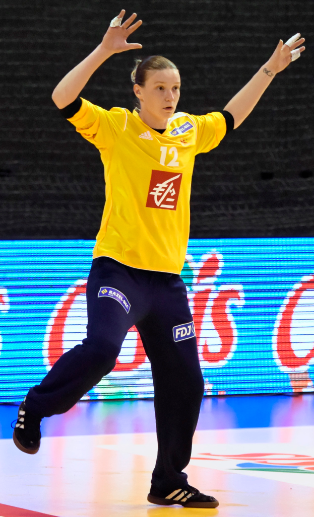 Eine Frau in einem gelben T-Shirt und schwarzen Hosen spielt Handball, trägt Schuhe und hat einen Bildschirm mit Text im Hintergrund.