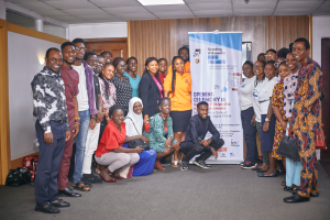 Eine Gruppe von Menschen, die vor einem Banner an einer Schulöffnung in Lagos State posieren, wobei einige stehen und andere im Schneidersitz sitzen, Taschen auf dem Boden, eine Tür, ein Schild, ein Foto auf der Wand und eine Decke mit Deckenleuchten und einer Überwachungskamera.