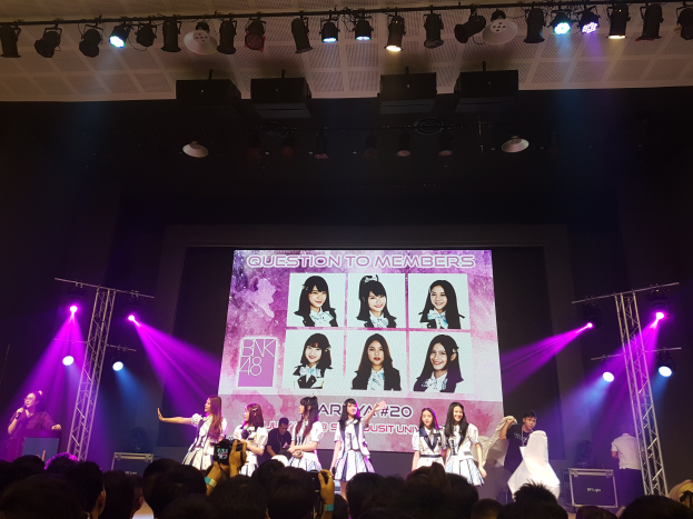 Eine Gruppe von Menschen, die auf einer hell erleuchteten Bühne performen, mit einer Menge darunter und einem Bildschirm im Hintergrund, der Bilder von BK48 anzeigt.