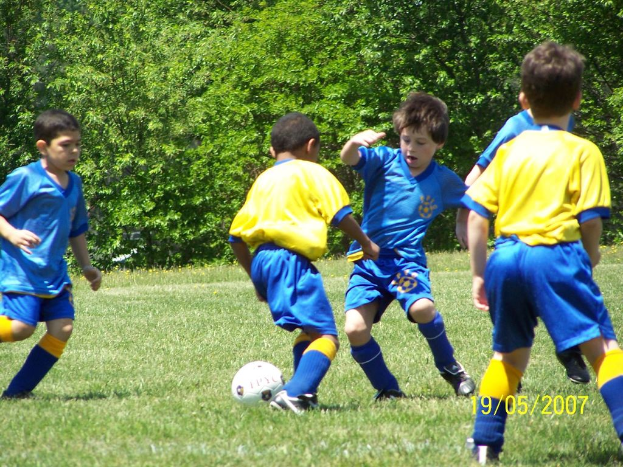 Kinder beim Fußballspielen auf einem grünen Rasen mit Bäumen im Hintergrund und einem Datum in der rechten unteren Ecke.