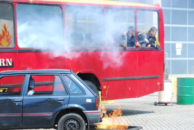 Ein roter Doppeldeckerbus mit Rauch, der aus ihm herauskommt, mit drei sichtbaren Passagieren, neben einem Auto geparkt, vor einem Gebäude mit Glasfenstern und einem Fass auf der rechten Seite.
