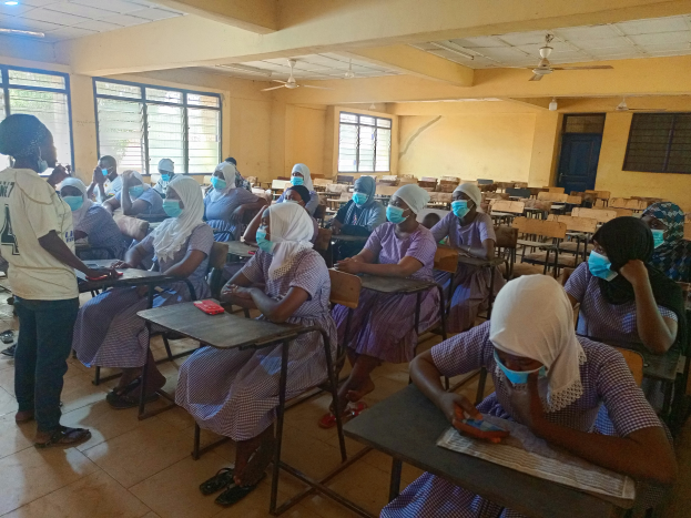 Ein Klassenzimmer in Nigeria mit Schülern, die Gesichtsmasken tragen und auf Bänken sitzen oder stehen, mit Büchern und Gegenständen auf den Tischen, Fenstern, einer Tür, einer Wand, einer Decke mit Lampen und Ventilatoren im Hintergrund.