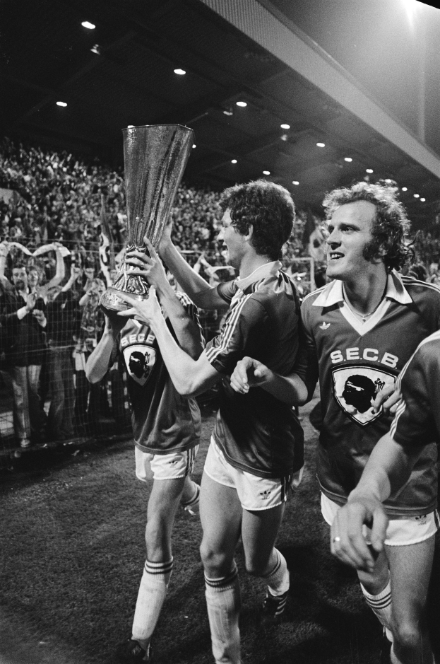 Schwarzes Bild von Fußballspielern, die mit einem Pokal auf einem Feld feiern, umgeben von einer Menge hinter einer Absperrung unter Stadionbeleuchtung.