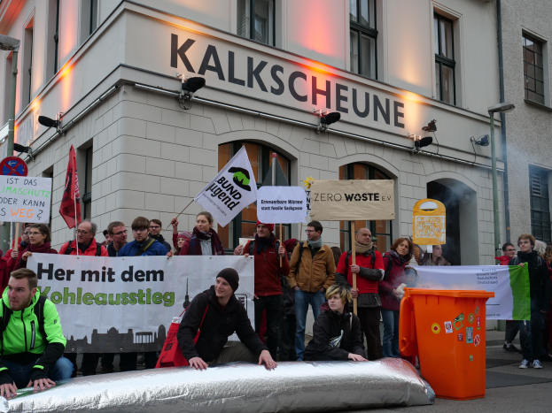 Eine Gruppe von Menschen mit Transparenten und Plakaten steht vor einem Gebäude, mit zwei Personen im Vordergrund und einem Mülleimer rechts, während einer Demonstration in Deutschland.