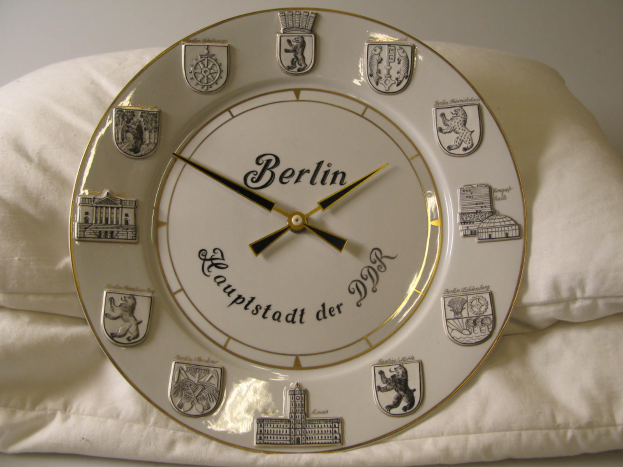 Eine Uhr mit dem Wort 'Berlin' darauf steht auf einem weißen Kissen vor einer Wand im Hintergrund.