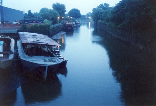 Viele Boote schwimmen neben einem Landgebiet mit geparkten Fahrzeugen, umgeben von Bäumen und einem Zaun um den Fluss.