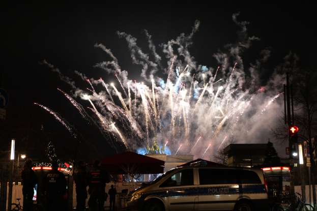 Ein Polizeiwagen vor einer Menschenmenge bei einer Silvesterfeier in Berlin geparkt, mit farbigen Feuerwerken, die den Himmel erhellen und die umliegenden Gebäude, Bäume und Schilder beleuchten.