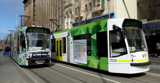 Zwei grüne und weiße Straßenbahnen auf einer Stadtstraße mit Fußgängern auf der linken Seite, Gebäuden, Bäumen, Pfählen, Drähten und einem klaren blauen Himmel im Hintergrund.