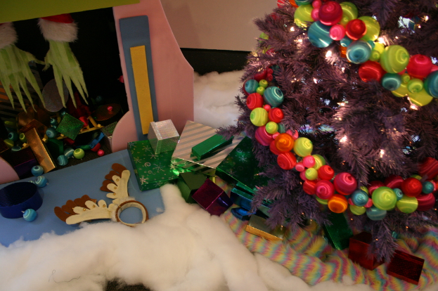 Eine Indoor-Szene mit einem Weihnachtsbaum mit Lichterkette und Dekorationen rechts, Geschenkboxen und Dekorationsgegenständen in der Mitte und einer Hütte mit Dekorationen und Watte oben links in der Ecke, mit einer Wand im Hintergrund und Watte und wollener Stoff unten.