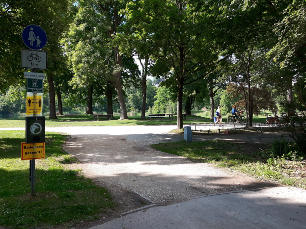 Ein Park mit einem asphaltierten Weg, der von Bäumen und Gras umgeben ist, Schilder an der linken Seite des Weges und Menschen, die im Hintergrund Fahrräder fahren.