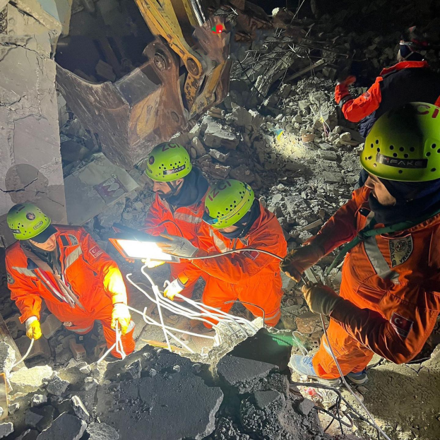Eine Gruppe von Menschen in orangefarbenen Uniformen und grünen Helmen bei der Arbeit an Schutt mit einem Kran im Hintergrund während einer Rettungsaktion.