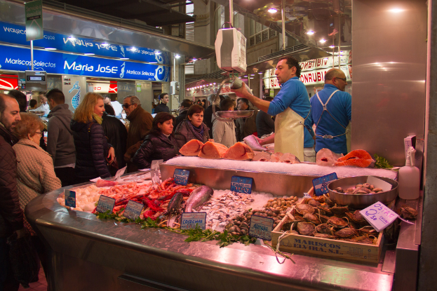 Eine Marktszene mit Menschen, die gehen und einige Fische schneiden, Fische und Bretter unten und Geschäfte, Bretter, Pfosten und Lichter im Hintergrund.