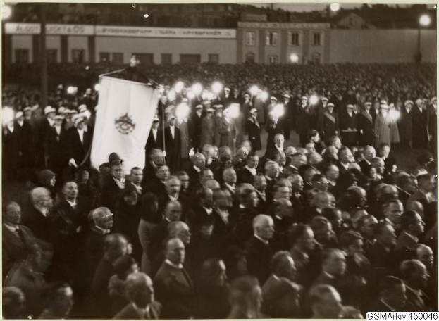 Schwarzes und weißes Foto einer großen Menschenmenge vor einem Gebäude, mit einer Person, die eine Fahne hält, Pfosten und Lichter im Hintergrund und Text unten, der 'Gustav Klimts Rede an der Berliner Mauer' lautet.