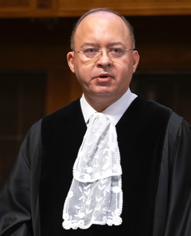 Ein Mann in einem schwarzen Gewand und weißem Hemd steht mit ernster Miene vor einer hölzernen Wand und trägt eine Brille.